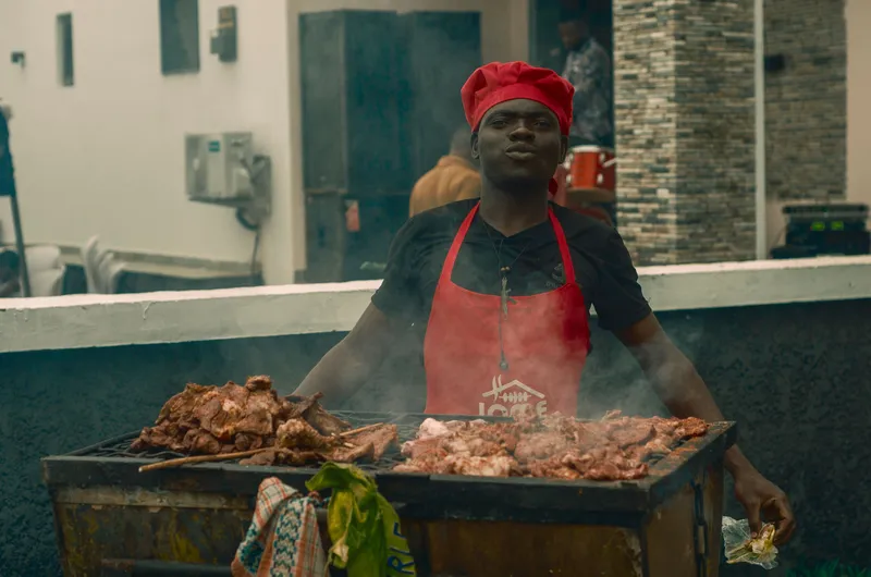 A street vendor wearing a red apron and hat grills meat outdoors, creating a smoky atmosphere.