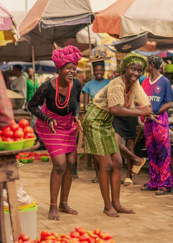 Two women in colorful traditional attire dance joyfully at a lively outdoor market.