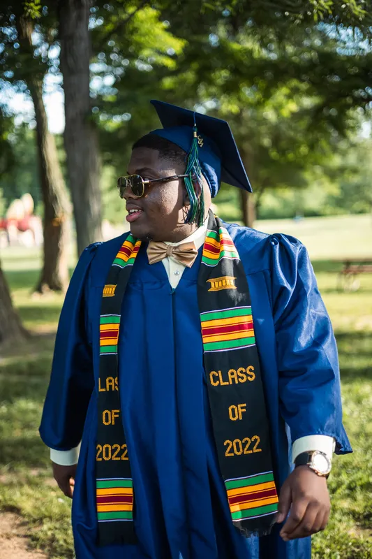 Smiling graduate in blue cap and gown with kente stole celebrating outdoors in a park.