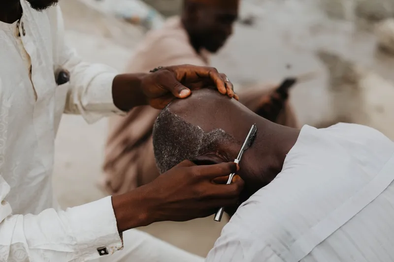 A barber meticulously shaves a customer's head outdoors, emphasizing traditional grooming.